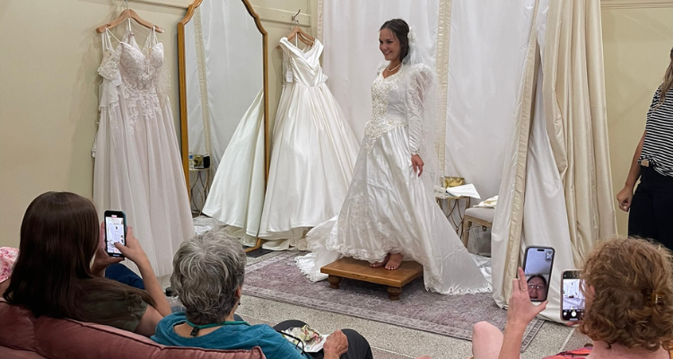 A bride-to-be smiles as she holds up a flowing tulle wedding dress against her body while surrounded by dresses in a bridal boutique, showcasing the excitement of wedding dress shopping.