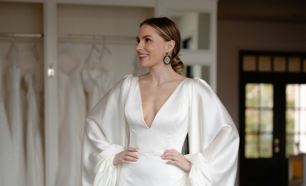 A bride-to-be smiles as she holds up a flowing tulle wedding dress against her body while surrounded by dresses in a bridal boutique, showcasing the excitement of wedding dress shopping.